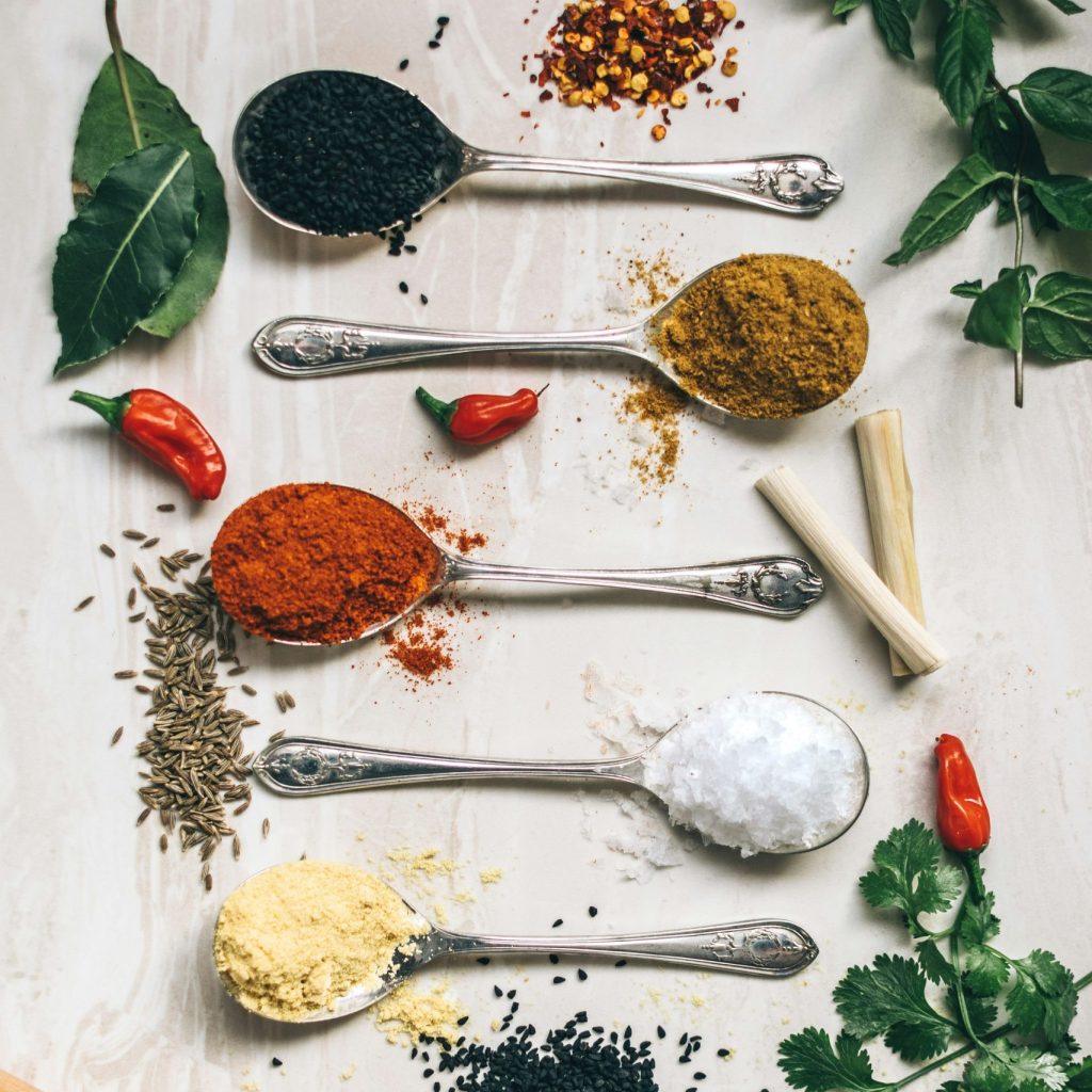 Assorted spices including turmeric, chili powder, black sesame seeds, cumin seeds, dried red chilies, and salt displayed with fresh herbs like cilantro and bay leaves on a white background.