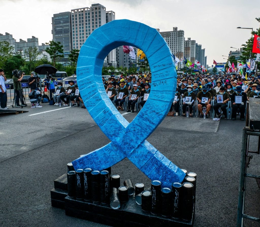 Blue awareness ribbon sculpture with candles at memorial event for victims, large crowd seated behind, cityscape in background, signs of remembrance and solidarity, LaNolaMom image.