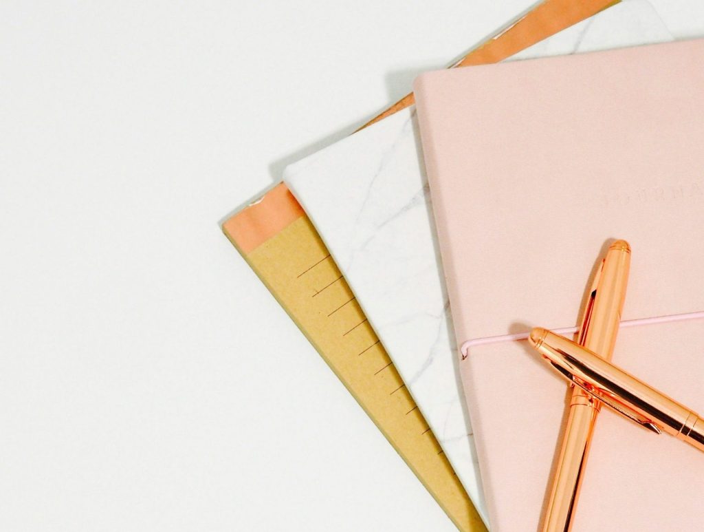 Elegant pink hardcover journal with a rose gold pen on top, surrounded by additional notebooks with marble and pastel covers, perfect for writing, journaling, or planner organization.