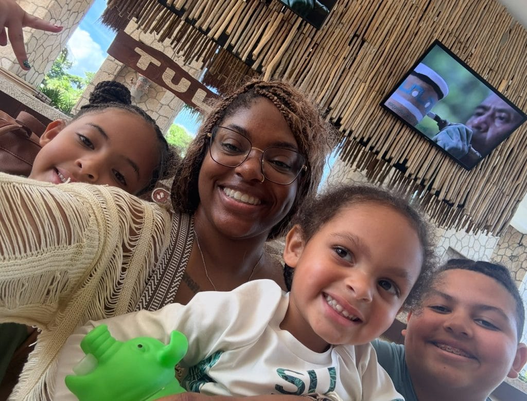 Woman smiling with three children at restaurant, family enjoying meal and taking selfie, tropical decor, TV in background, casual family outing, LaNolaMom lifestyle content, joyful family moment, outdoor dining experience, diverse family group, happy family photo, tropical restaurant ambiance.