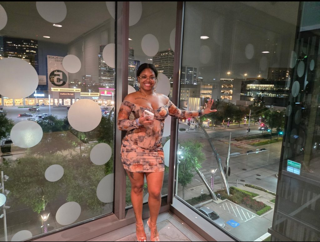 LaNolaMom standing in city view window, smiling...symbolizing hope, confidence, and storytelling after loss.