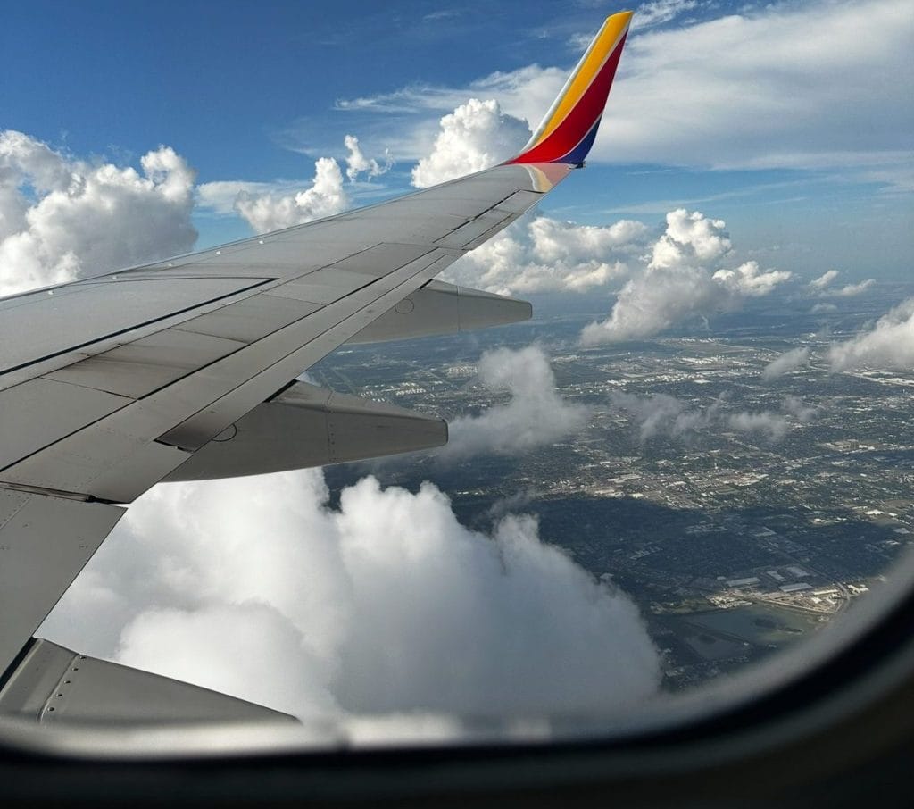 Wing of an airplane flying high above the clouds with a view of the sky and landscape below.